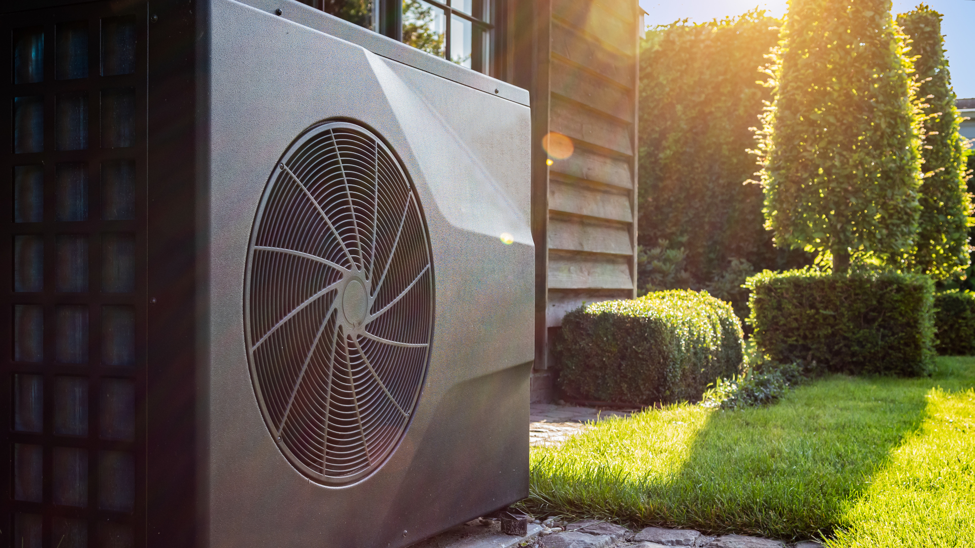 Close-up of black full inverter heat pump outside in the garden, near wooden pool house on a sunny day. Lens flare on the image.