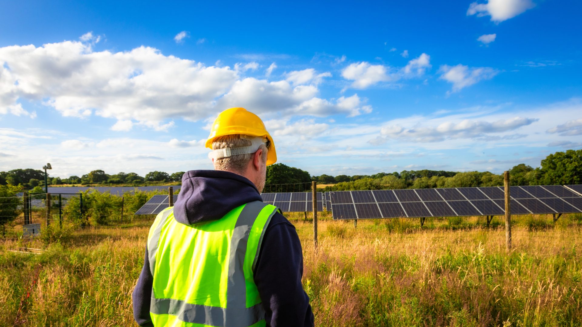 A mid adult male maintenance engineer dressed in reflective vest and hardhat at the solar farm. Banks of solar panels are in the background.